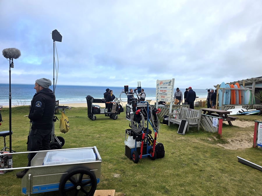 Film crew sets up equipment on a grassy area overlooking a beach with surfboards nearby