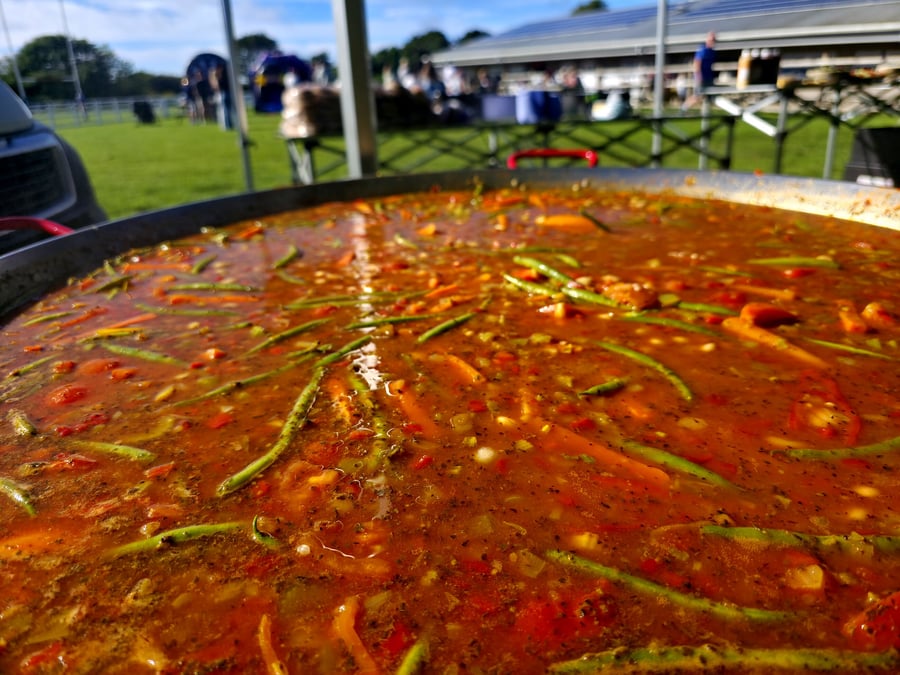A large pan of paella cooking outdoors with vegetables and a vibrant sauce