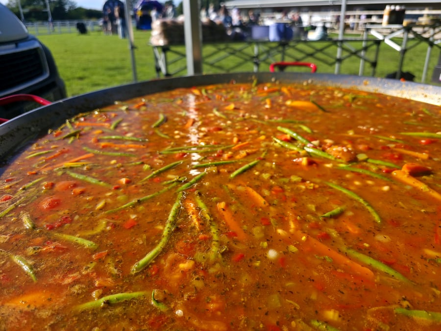 A large pan of paella cooking outdoors at an event with people in the background