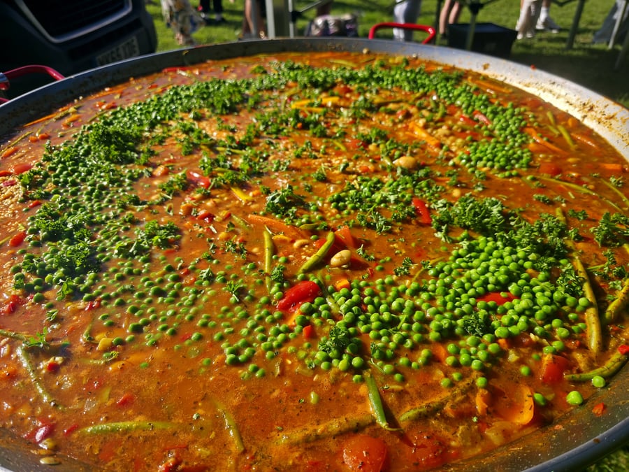 A giant pan of paella with peas, peppers, and herbs, cooked outdoors