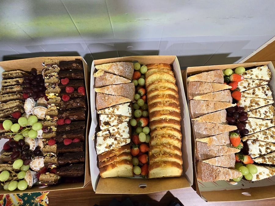 Three catering boxes filled with assorted sweet traybakes, fruit, and bread