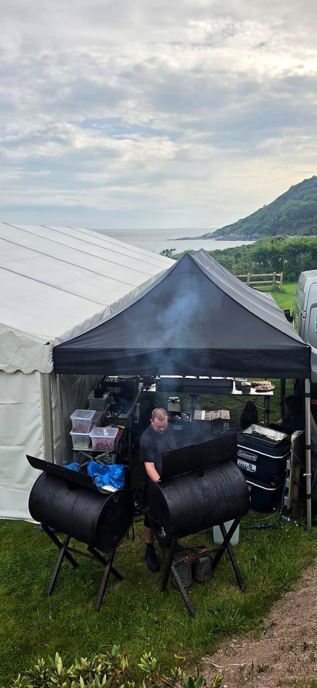 Catering team cooking outdoors with smoke rising from barrel BBQs by the coast