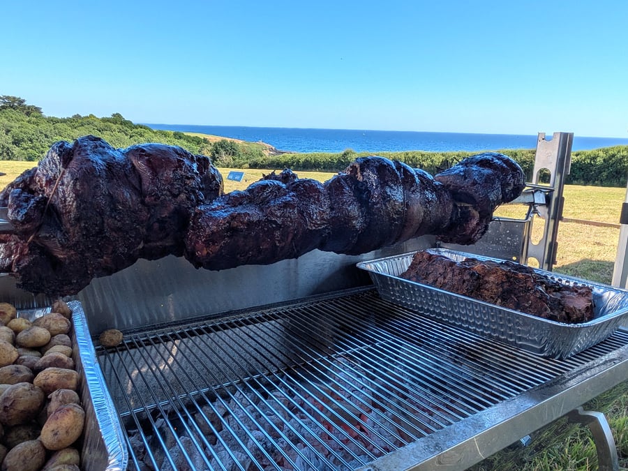 A large hog roast is cooking outdoors with potatoes and the sea in the background