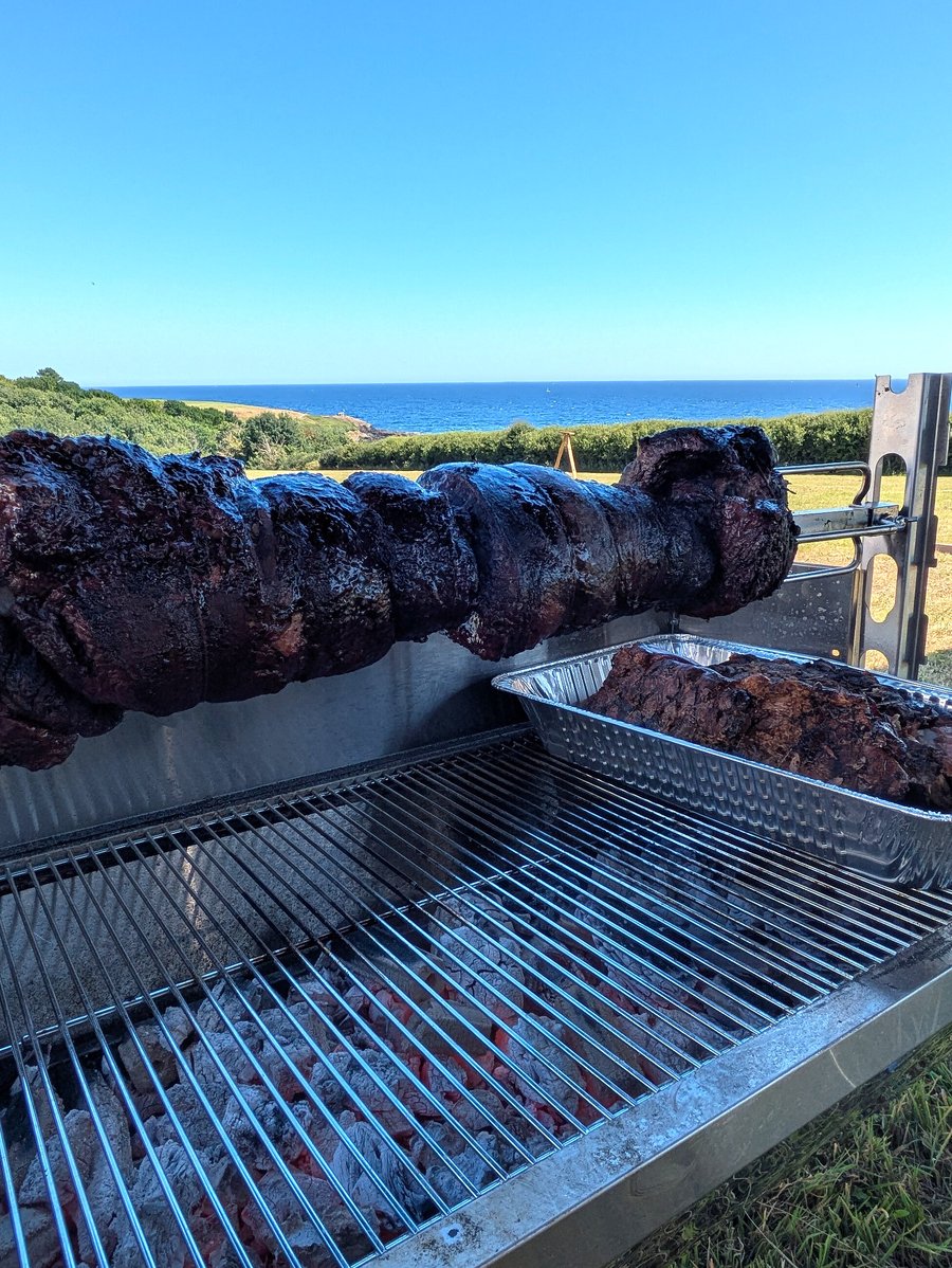 A large spit-roasted meat cooks outdoors with a beautiful sea view