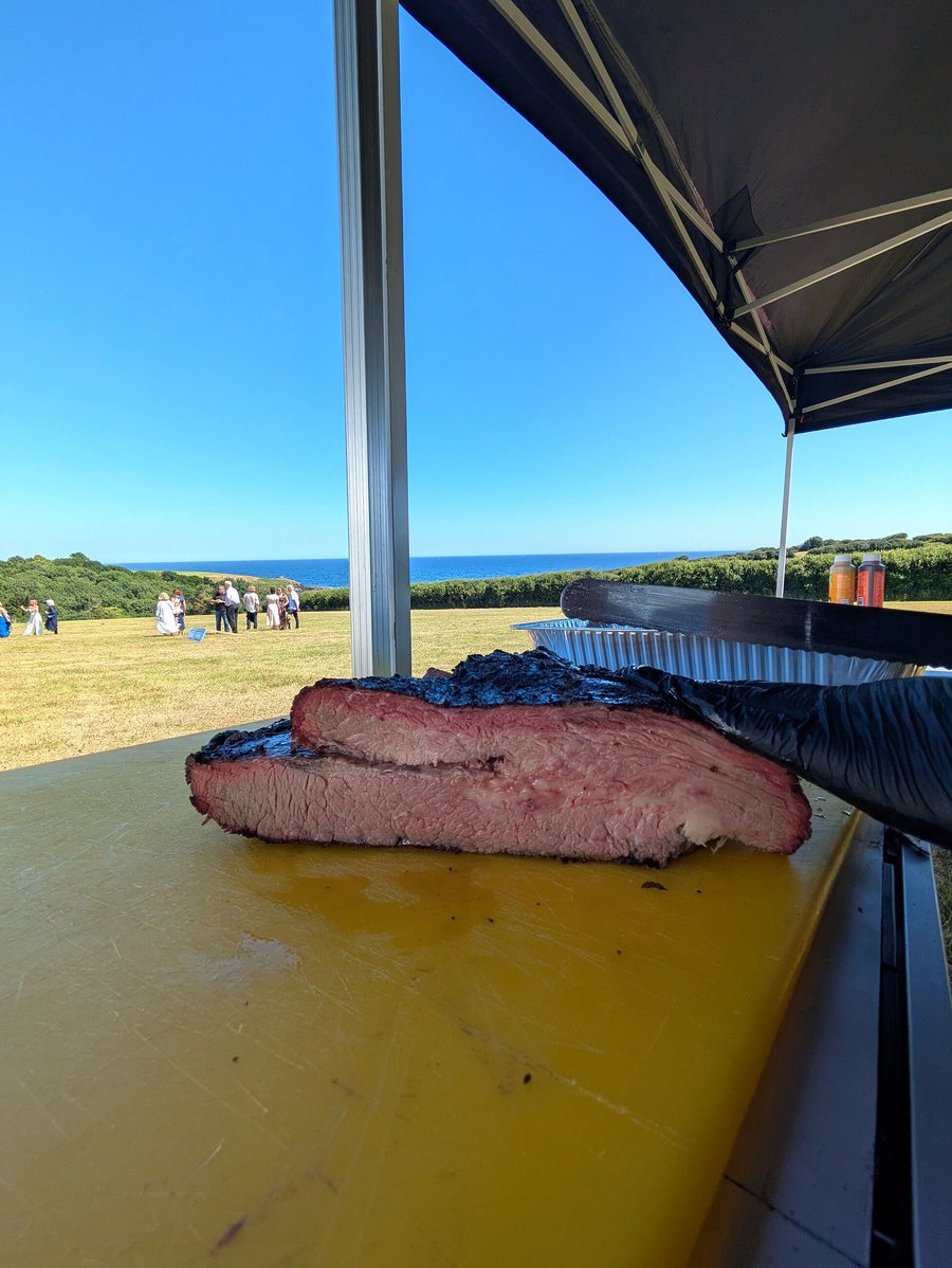 A large, perfectly cooked brisket sits on a yellow cutting board with the ocean in the background