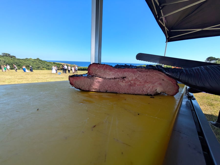 A large, perfectly cooked brisket is carved outdoors with a beautiful coastal view