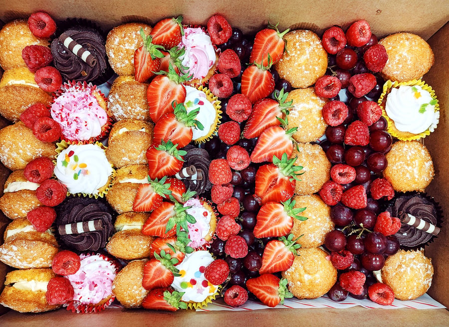 A box filled with assorted mini cupcakes, fresh strawberries, raspberries, and grapes