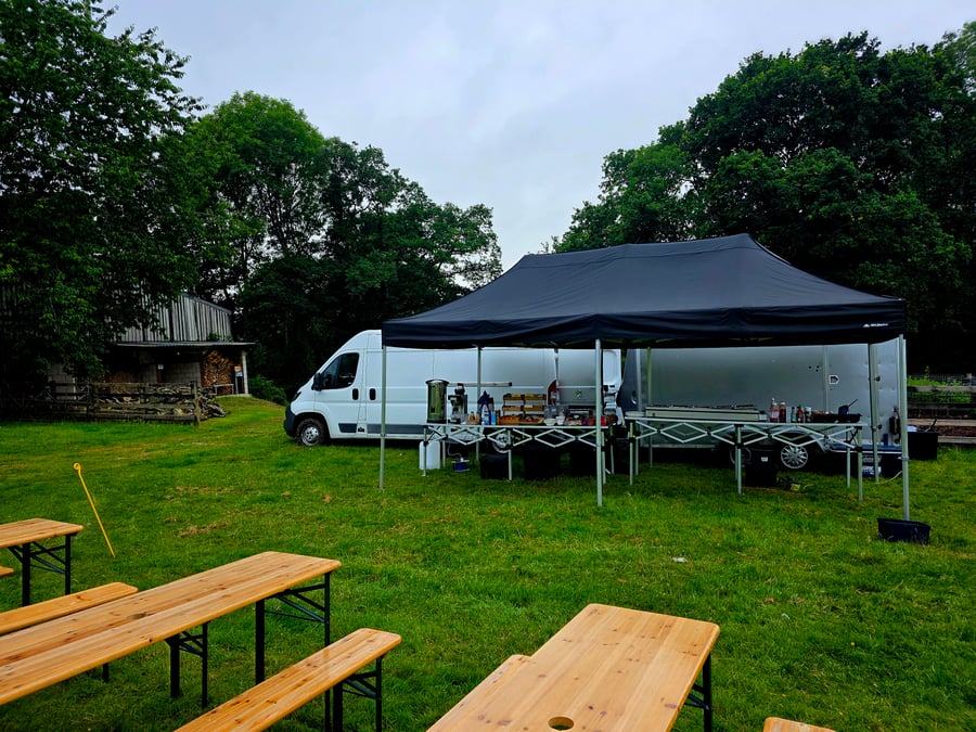 A catering setup with a gazebo, van, trailer, and wooden picnic benches in a grassy field, ready for an outdoor event