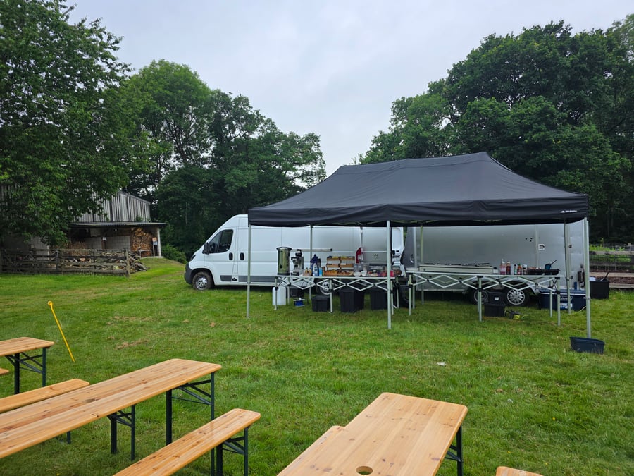 A catering setup with a gazebo, van, trailer, and picnic benches in a grassy field, ready for an outdoor event