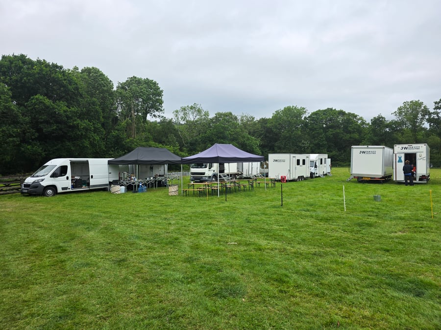 An outdoor catering setup with several marquees, vans, and tables on a grassy field, ready to serve a film crew