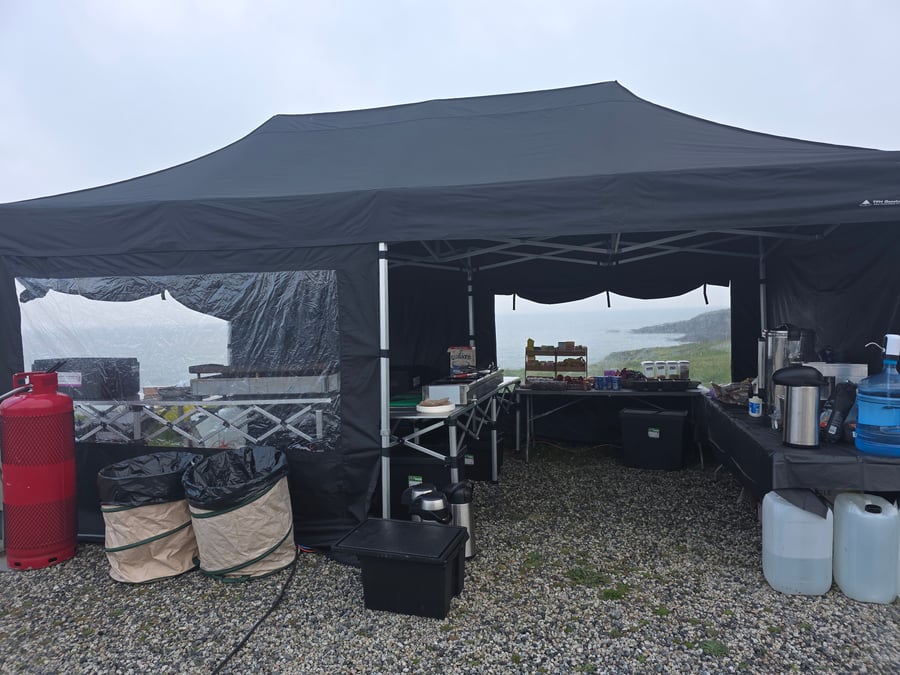 A black gazebo set up for an event, with catering equipment and a view of the coastline in the background