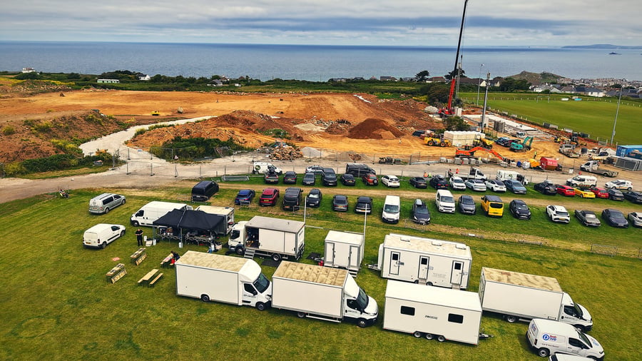 An aerial view of a film production unit base with catering tents and vehicles on a grassy field overlooking the sea