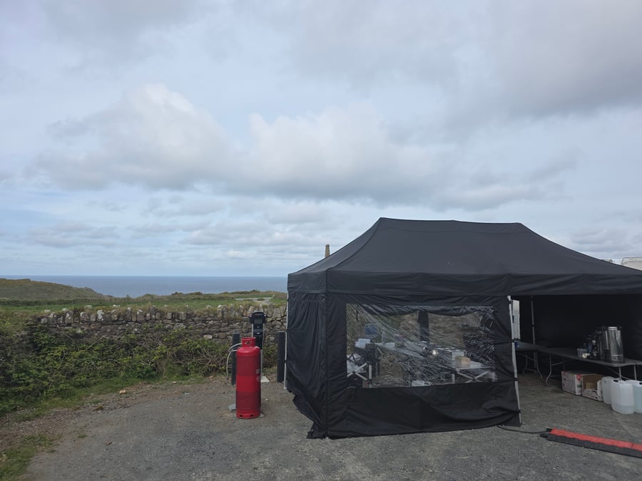 A black catering gazebo is set up on a gravel pitch with a stone wall and the sea in the background under a cloudy sky