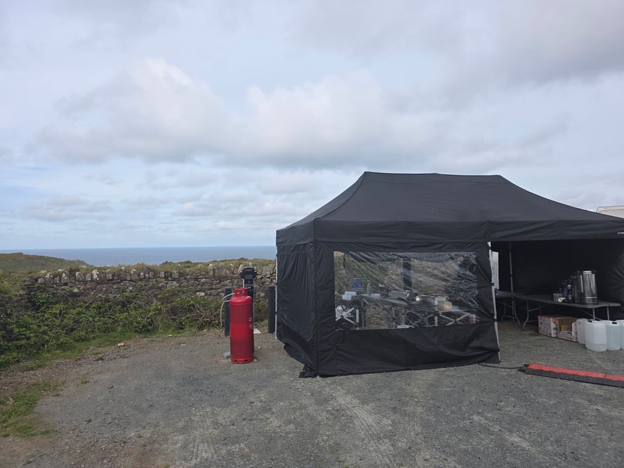 A black catering gazebo is set up on a gravel area next to a stone wall, with the sea and cloudy sky in the background