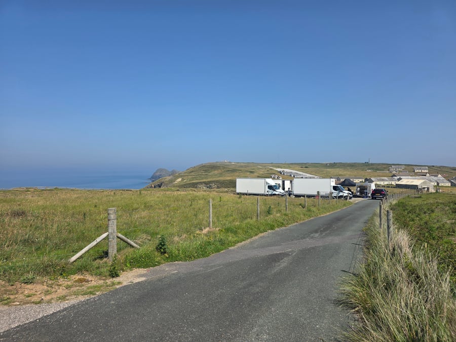 A film or TV production unit base with catering vehicles and trailers set up in a coastal field on a sunny day