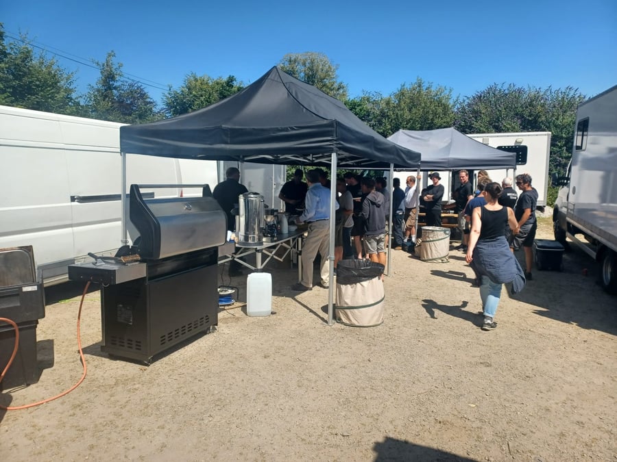 A busy film crew unit base with catering gazebos, a BBQ, and a queue of people waiting for food