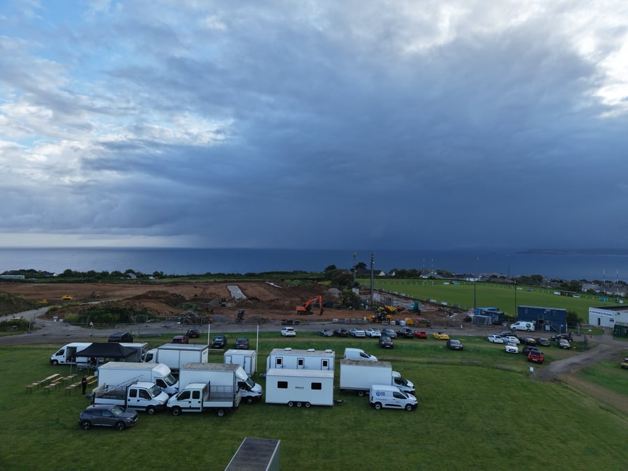 Aerial of a film unit base on the Cornish coast at dawn under a dramatic sky — the catering gazebo, box trucks and welfare trailers set up above the sea