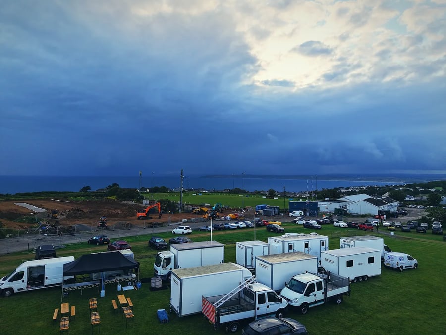 Aerial view of a film unit base above the Cornish coast — the catering gazebo, box trucks and welfare trailers set up under a dramatic sky