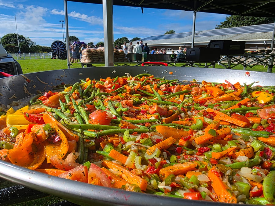 A vibrant mix of fresh vegetables, including green beans, carrots, and tomatoes, cooking in a giant paella pan at an outdoor event