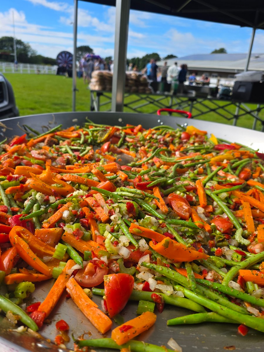 A vibrant mix of fresh vegetables, including carrots, green beans, and peppers, cooking in a giant paella pan at an outdoor event