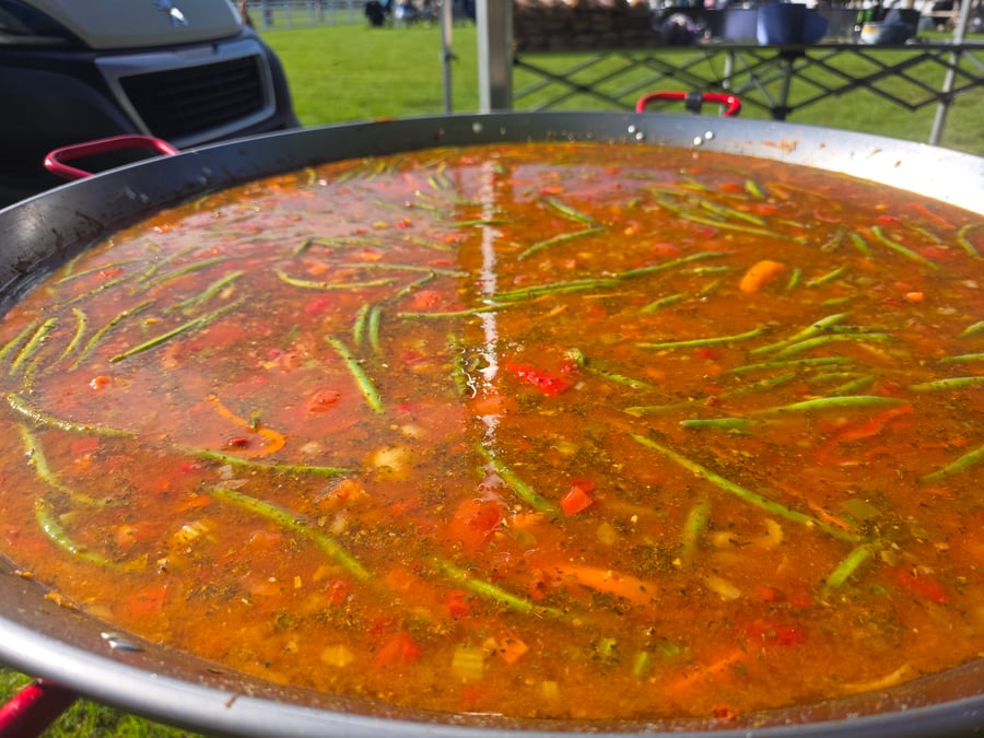 A vibrant vegetable paella simmers in a giant pan, ready to be served at an outdoor event
