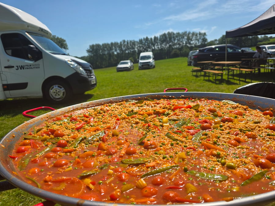 A giant paella pan cooking outdoors at a film and TV production unit base, with crew vehicles and marquees in the background