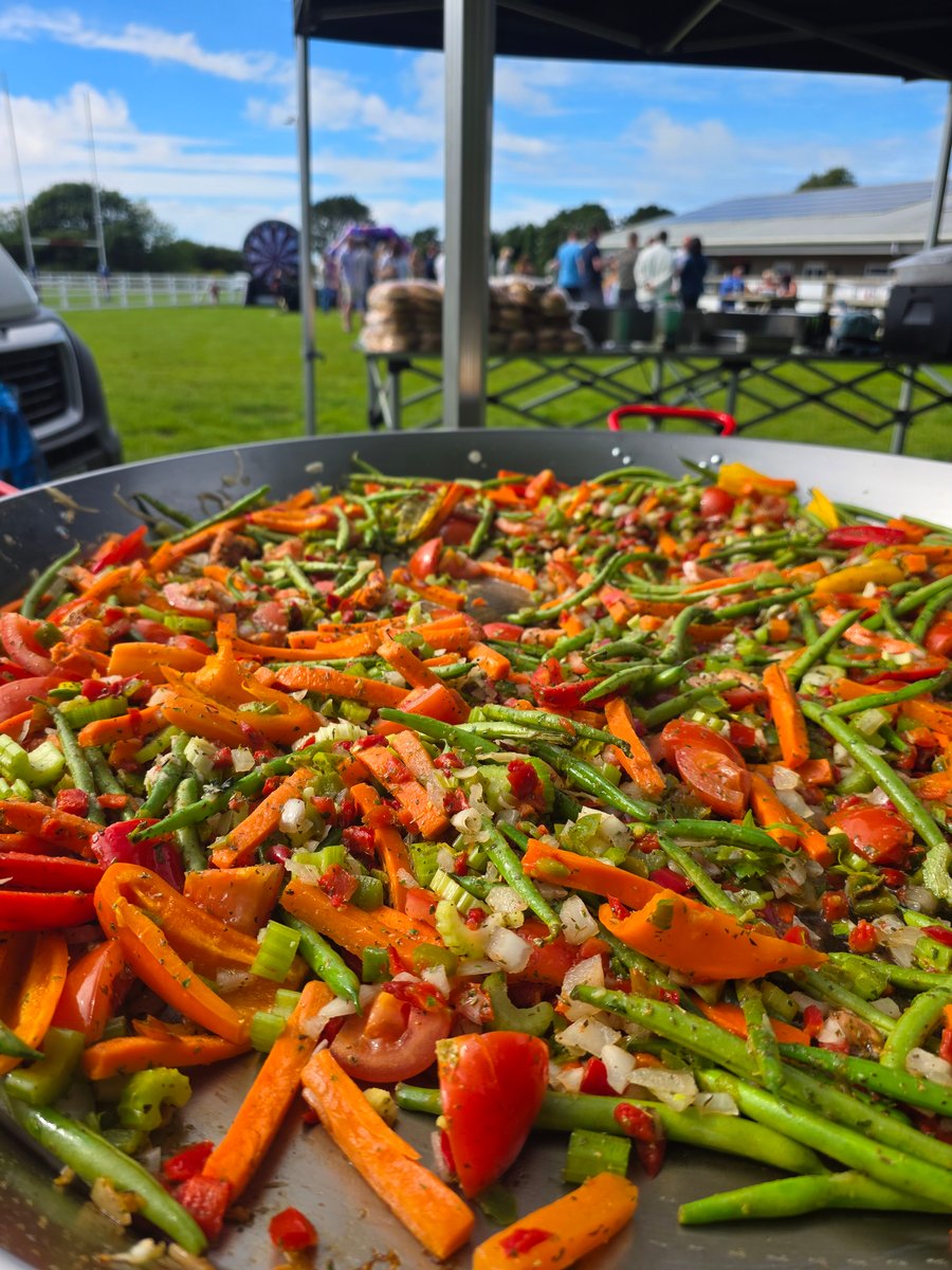 A vibrant giant paella pan filled with colourful vegetables, cooking outdoors at a lively event under a bright blue sky