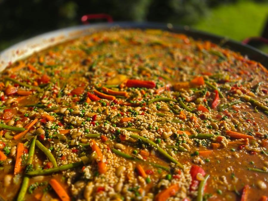 A vibrant giant paella pan filled with rice, green beans, carrots, and peas, cooking outdoors under natural light