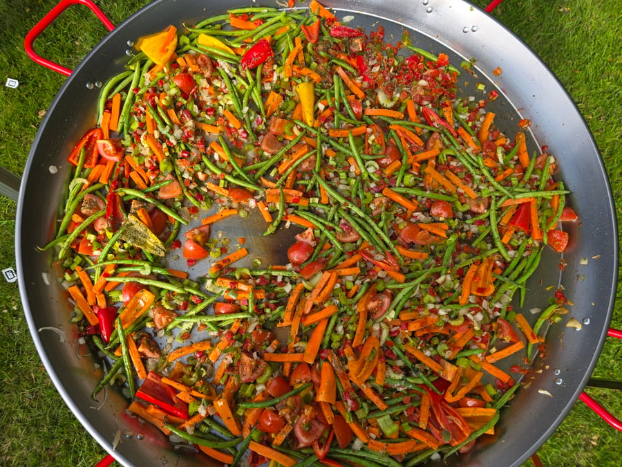 A vibrant mix of chopped vegetables, including green beans, carrots, and peppers, cooking in a giant paella pan outdoors