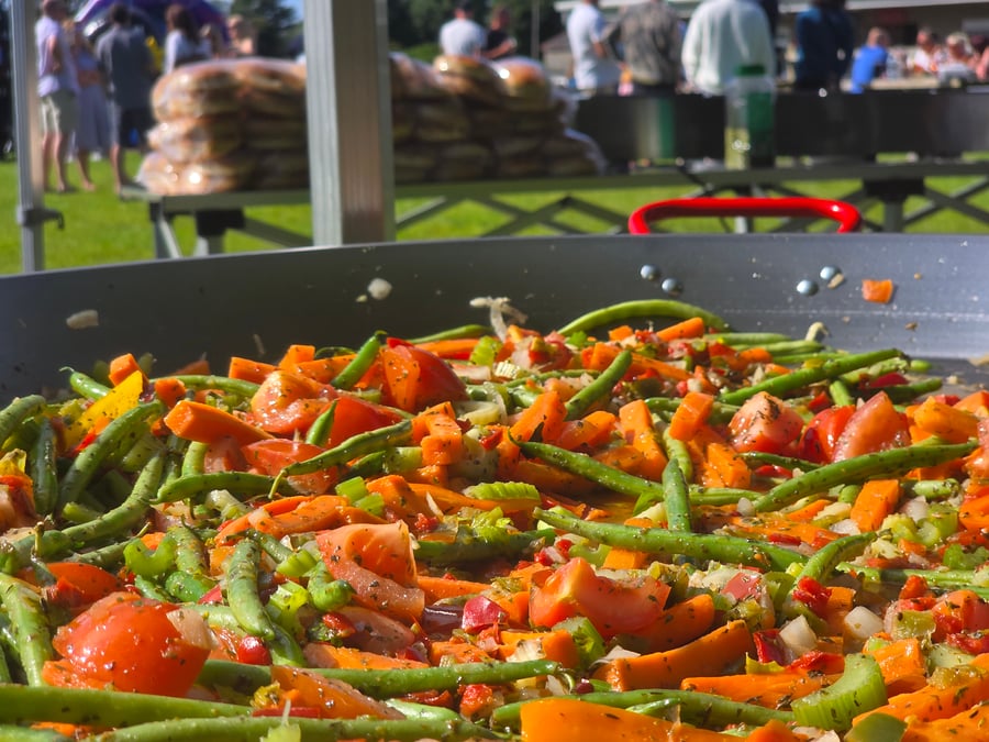 Freshly chopped vegetables, including green beans, carrots, and tomatoes, cooking in a giant paella pan at an outdoor event