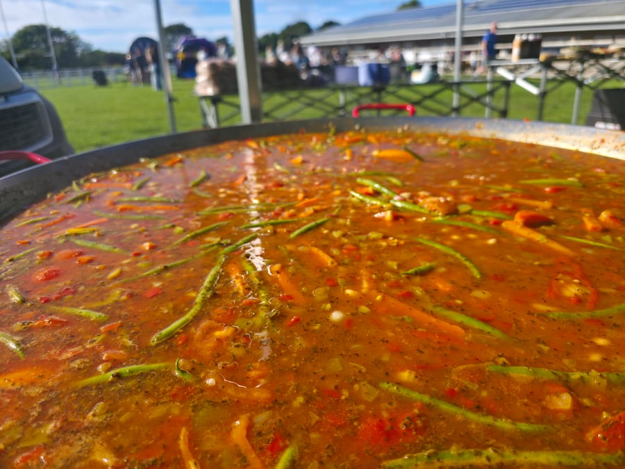 A vibrant giant paella pan filled with a rich, colourful stew of vegetables, cooking outdoors at an event