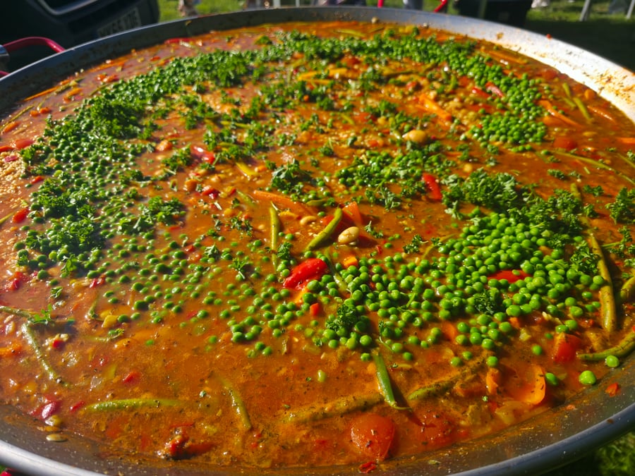 A vibrant giant paella pan filled with a colourful vegetable and rice dish, garnished with fresh peas and parsley