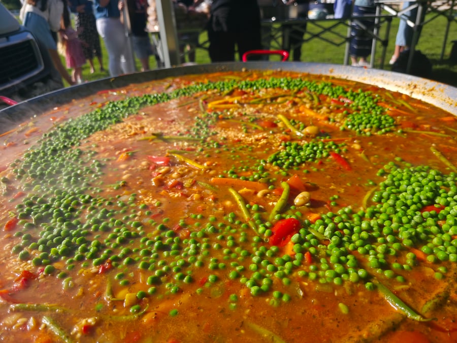 A vibrant giant paella pan brimming with rice, peas, and vegetables, cooking outdoors at a lively event