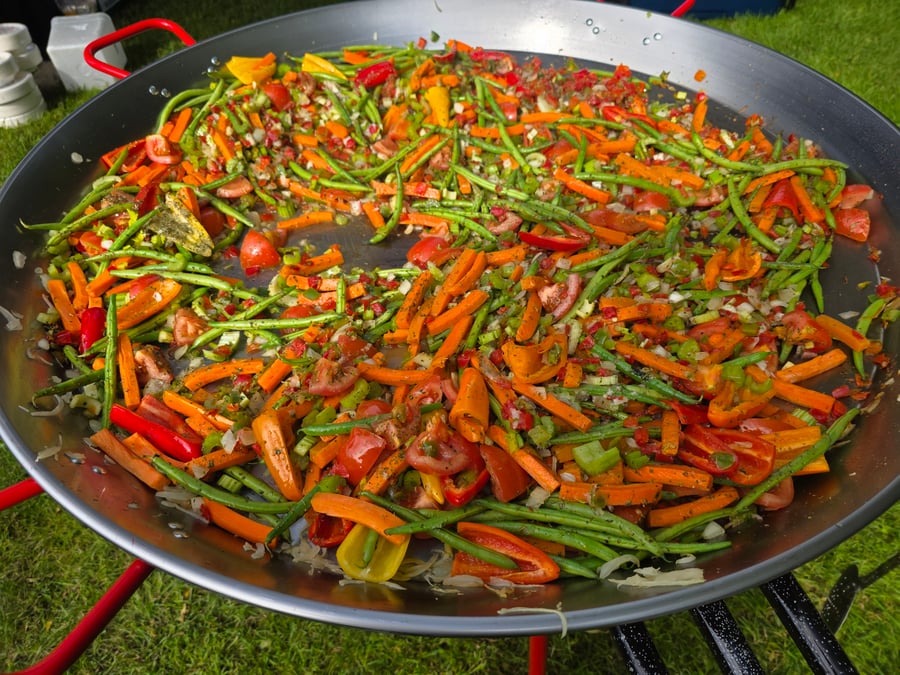 A vibrant mix of chopped vegetables, including carrots, green beans, and peppers, cooking in a giant paella pan outdoors