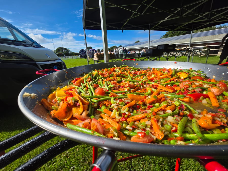 A vibrant giant paella pan filled with colourful vegetables, cooking outdoors under a blue sky at an event