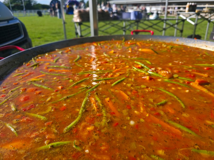 A giant paella pan filled with a vibrant vegetable and rice dish, cooking outdoors at an event