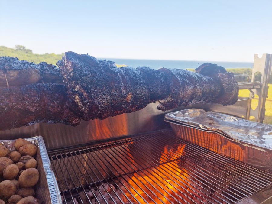 A hog roast slowly cooking over an open fire with roasted potatoes, set against a coastal backdrop