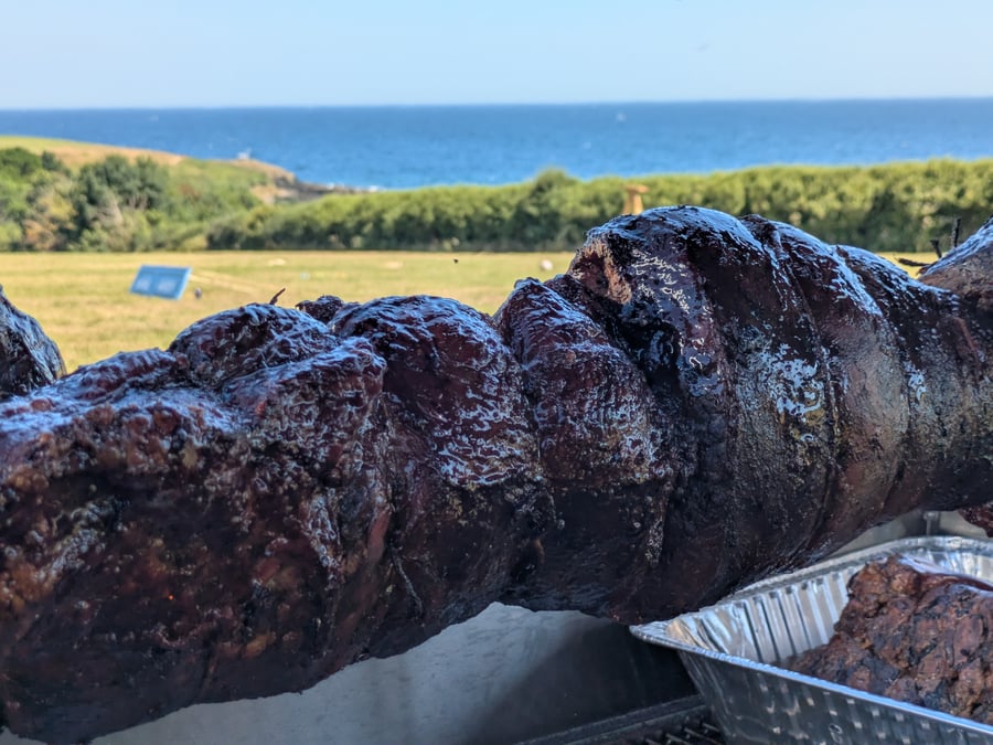 A whole hog roasts on a spit with a stunning coastal view in the background, ready to be carved for guests