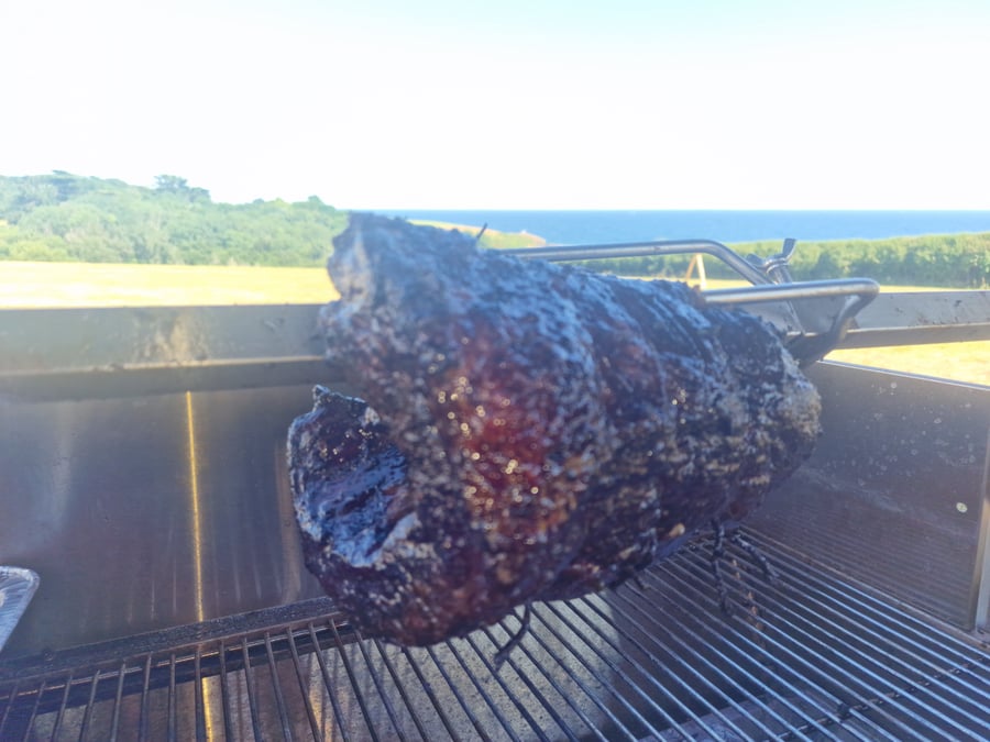 A hog roast cooking on a spit with the sea and green landscape in the background on a sunny day
