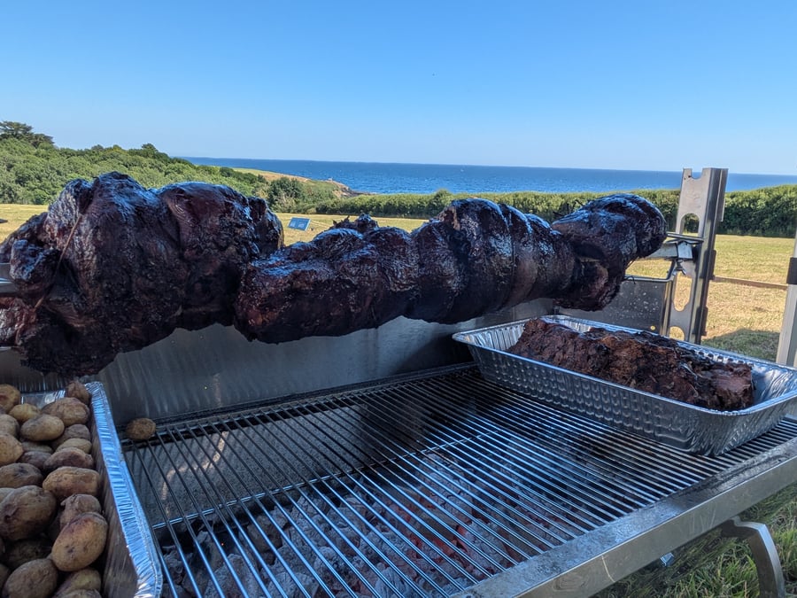 A hog roast cooking on a spit over hot coals, with roasted potatoes and a stunning coastal view in the background