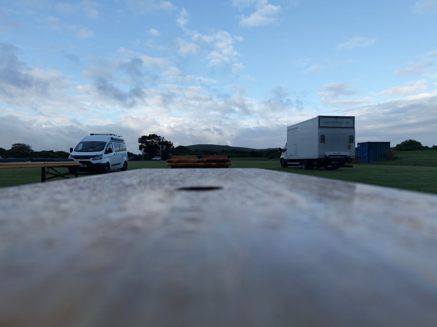 A film or TV production unit base with catering vehicles and a picnic bench on a grassy field under a cloudy sky