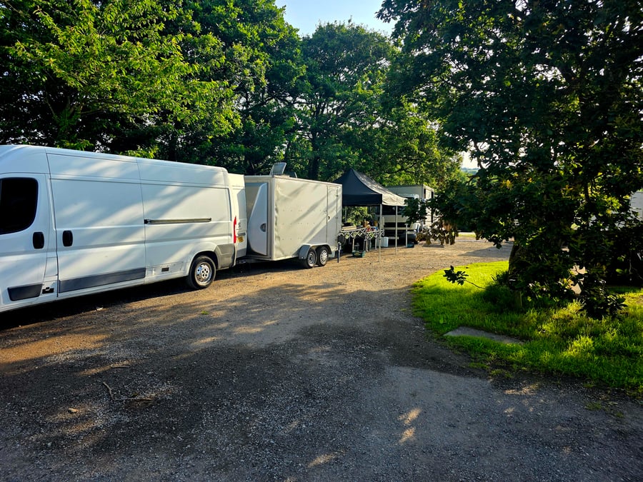 A white catering van and trailer parked on a gravel track at a film or TV production unit base, with a black gazebo and trees in the