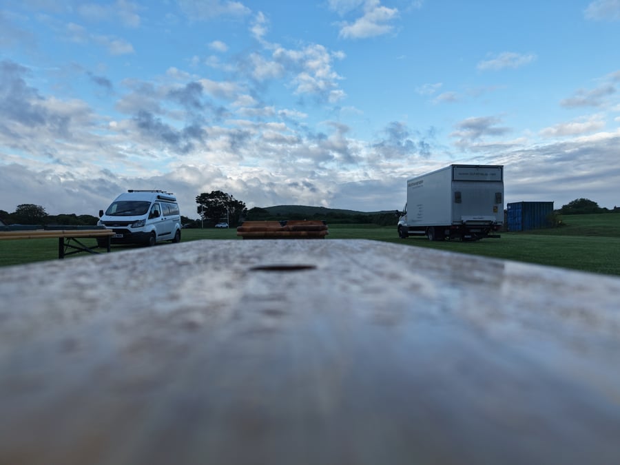 A film or TV production unit base with catering vehicles and a picnic bench under a cloudy sky