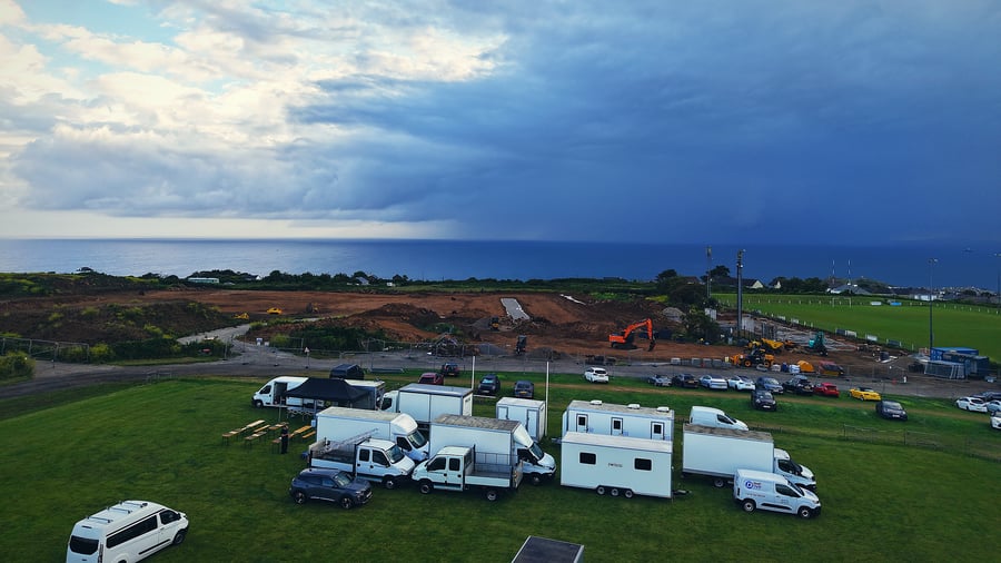 An aerial view of a film or TV production unit base with catering vehicles and welfare units, set against a dramatic coastal backdrop