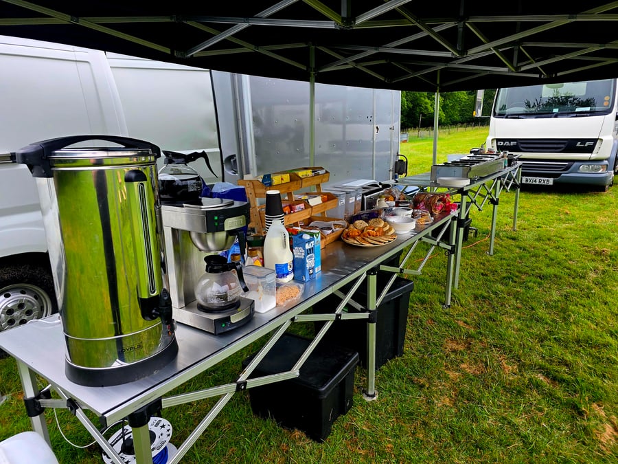 A breakfast buffet with hot drinks is set up under a gazebo at a film or TV production unit base