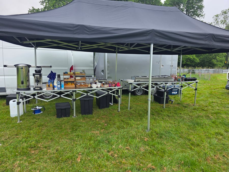 A catering setup under a black gazebo, featuring hot drinks, pastries, and cooking equipment, ready to serve a film or TV crew