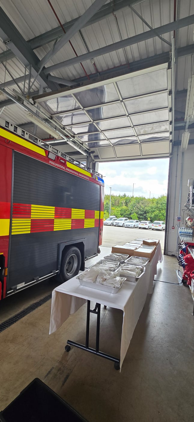 A table laden with individually packaged meals for welfare catering, set up inside a fire station next to a fire engine
