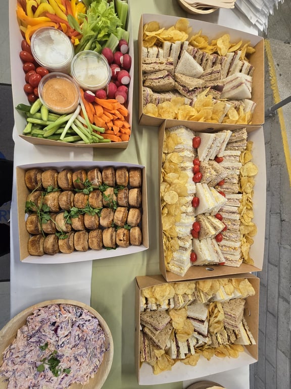 A December buffet laid out on a long table: boxes of sandwiches with crisps, sausage rolls, crudités with dips and a bowl of coleslaw