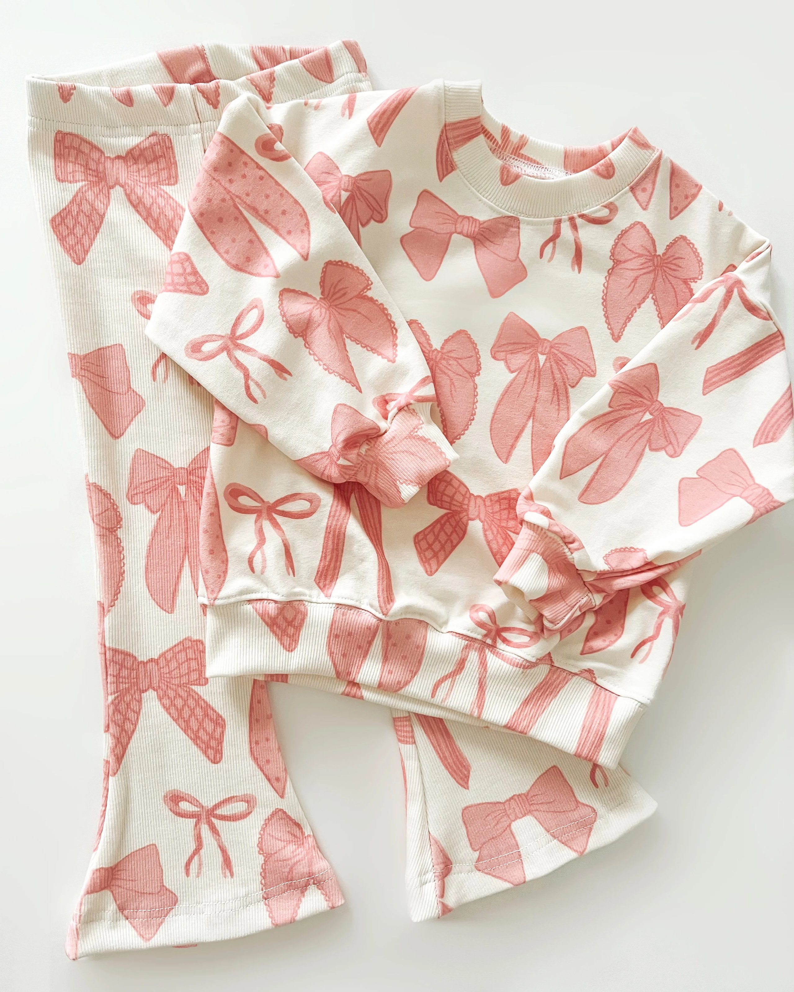 A small child’s two-piece set with a long-sleeved sweatshirt and flared-leg pants lays on a white background. The print on the set features pink hairbows in different styles on a cream background. 