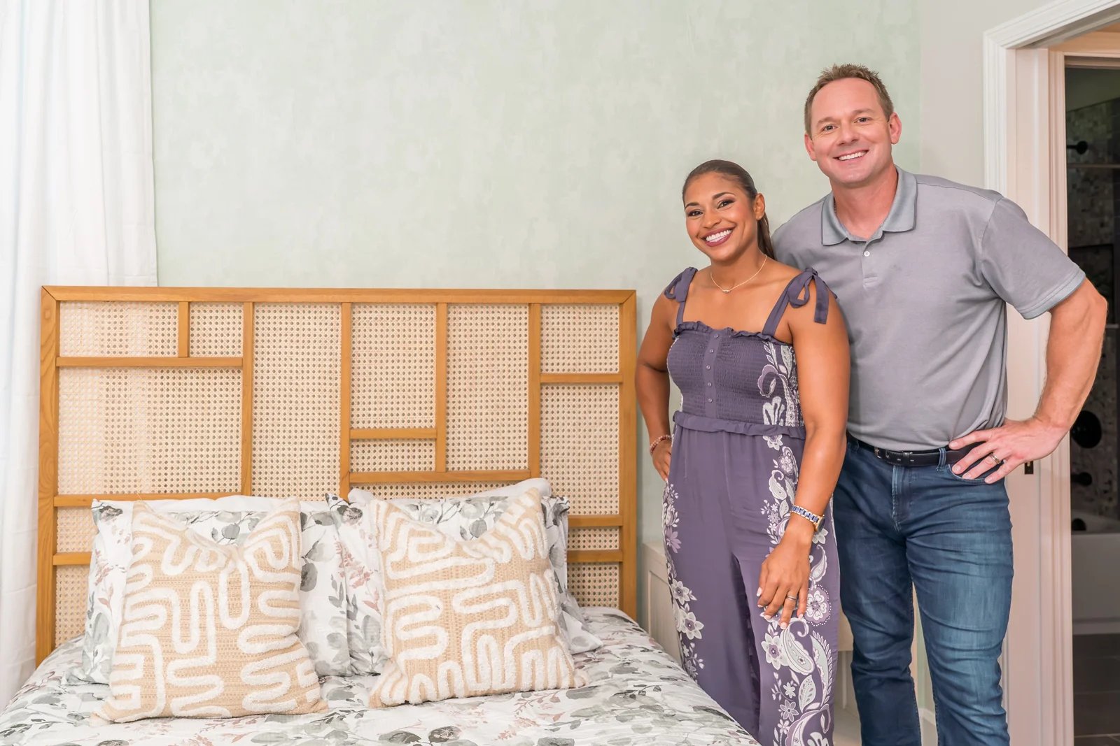 Brian and Mika, hosts of HGTV 100 Day Dream Home, stand by a bed with custom linens that coordinates with a wallpaper design from their collection at Little Cocalico. 